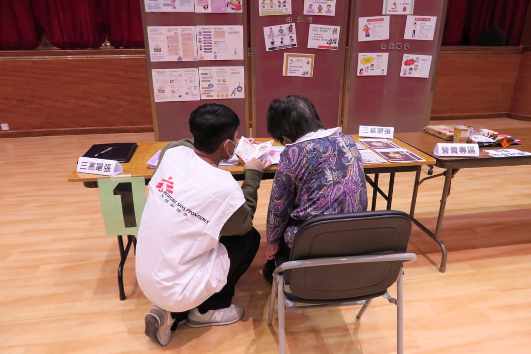 A man in an MSF vest kneels beside a woman sitting in a chair, helping her with papers on the table in front of them