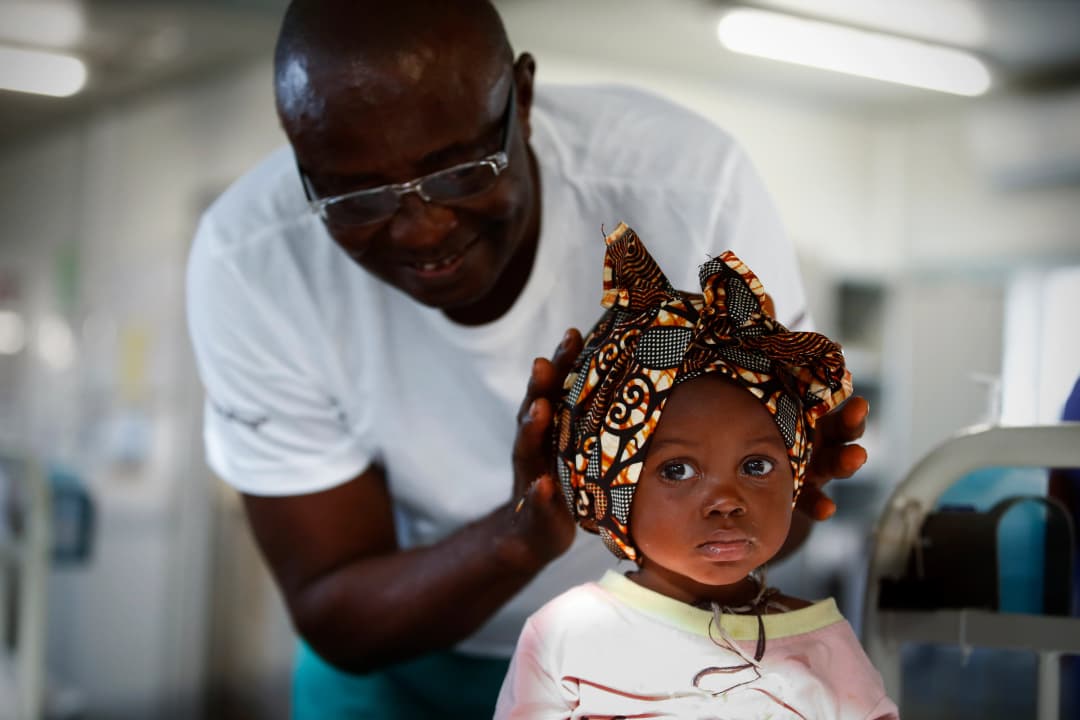 A one year old in a headscarf sucks her thumb and is attended by a healthcare provider. 